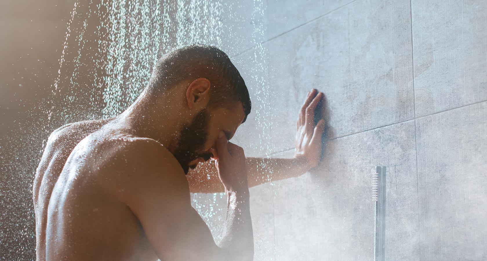 Mann steht unter laufender Dusche, stützt sich an der Wand ab und hält sich die Nase, während Wasser von oben herabfliesst.