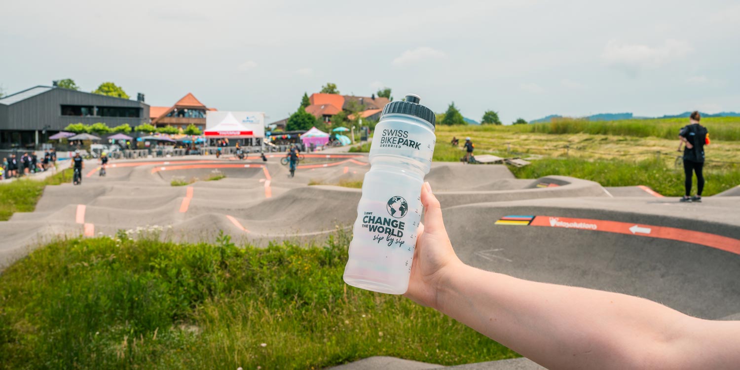 Hand hält Flasche, im Hintergrund ist ein Velo-Parcours.