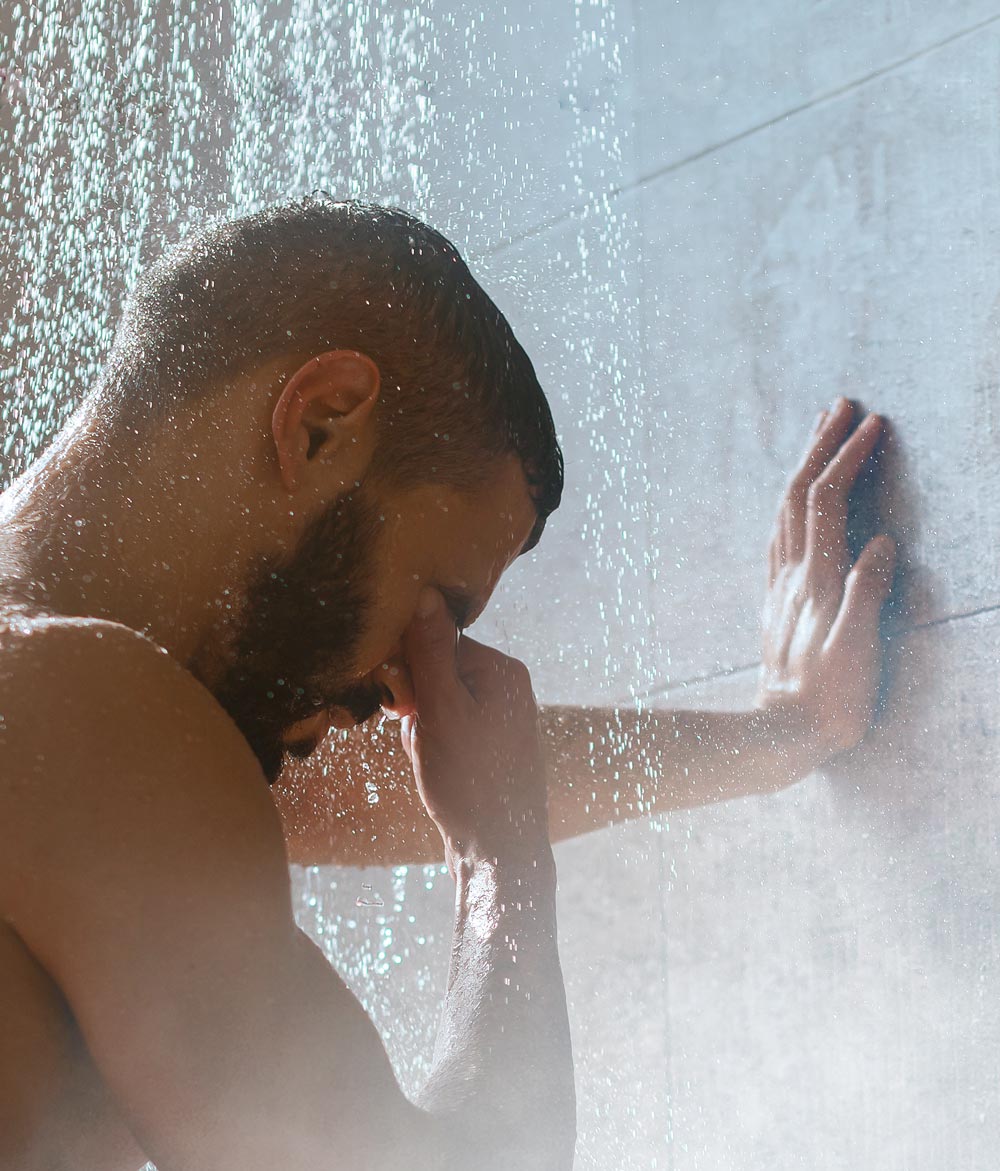 Un homme se tient sous une douche en fonctionnement, s’appuie contre le mur et se tient le nez pendant que l’eau coule d’en haut.