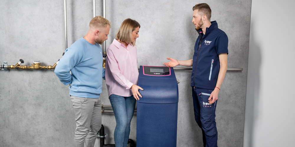 Un homme, une femme et un technicien de service BWT se tiennent à côté d’une installation d’adoucissement de l’eau.