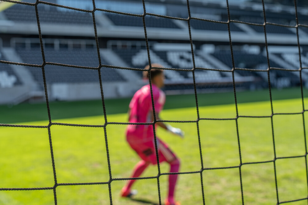 A blurred soccer goalkeeper in a bright pink uniform stands alertly in front of the goal, viewed through the netting.