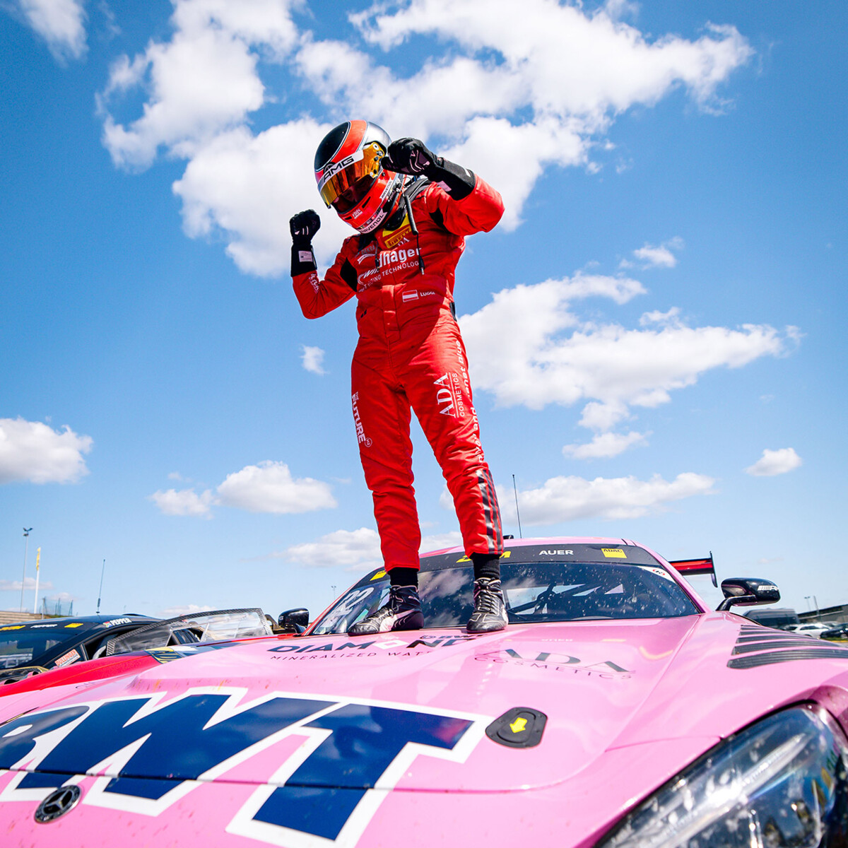 A professional race car driver stands triumphantly atop a pink BWT race car under a bright blue sky dotted with clouds.
