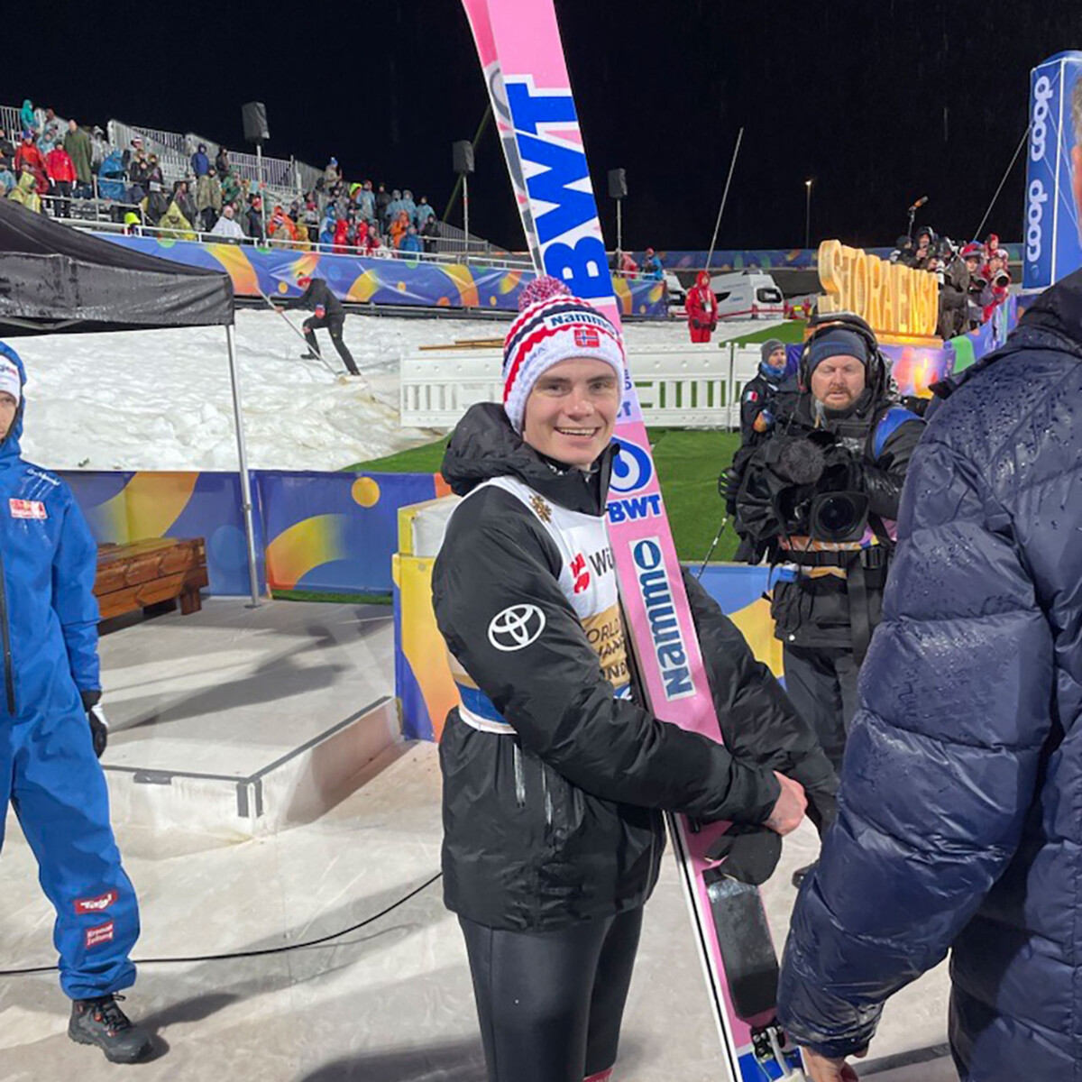 A smiling ski jumper poses with brightly pink colored skis at a professional winter sports event.