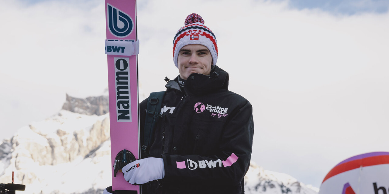 A professional ski jumper stands confidently against a snowy mountain backdrop.