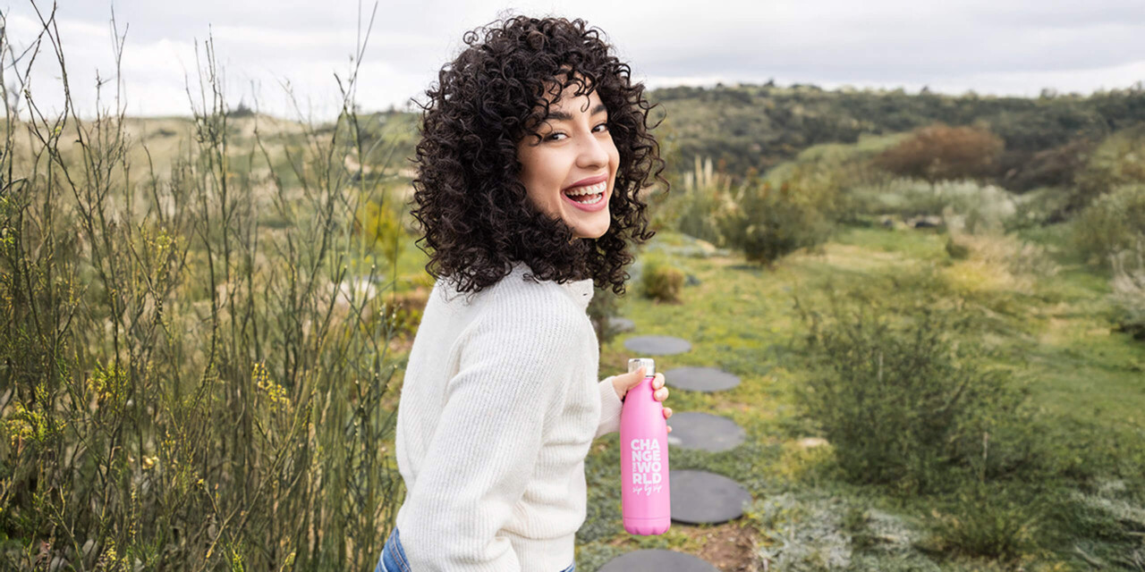 A young woman with curly hair and a white sweater holds a pink water bottle in her hand.