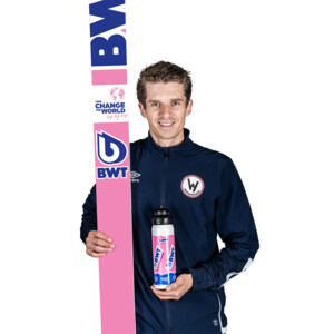 A male ski jumper stands confidently in a navy tracksuit, holding a pink BWT-branded ski and matching water bottle.
