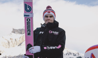 A professional ski jumper stands confidently against a snowy mountain backdrop.