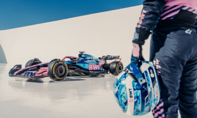 A Formula 1 race car in blue and pink BWT livery stands on a sleek, minimalist set. In the foreground, a race driver in matching suit holds a helmet.