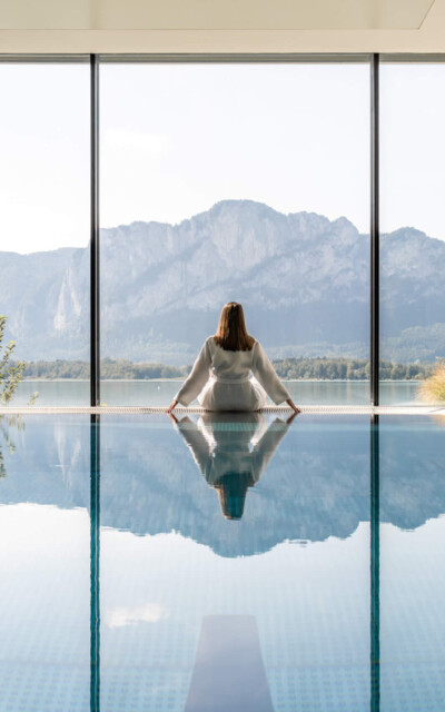 A woman sits relaxed at the edge of a modern indoor pool and enjoys the impressive view of a sweeping Alpine panorama.