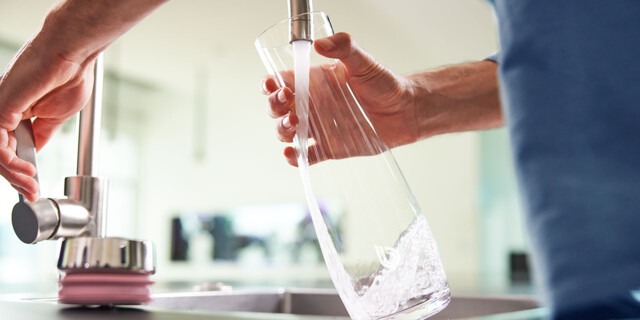 A person is filling a transparent glass carafe with fresh water from a modern kitchen faucet. 