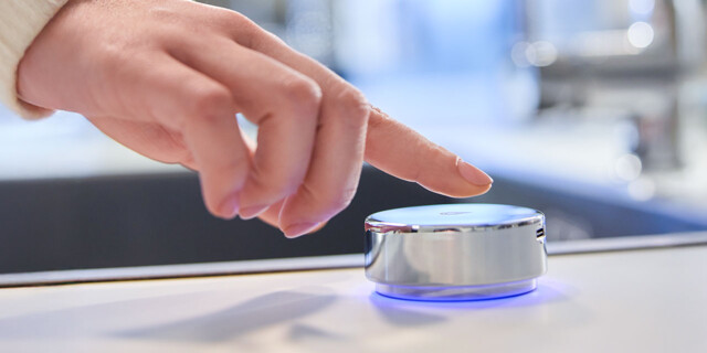A close-up of a person’s hand about to activate a sleek, circular smart water controller with a blue LED light.