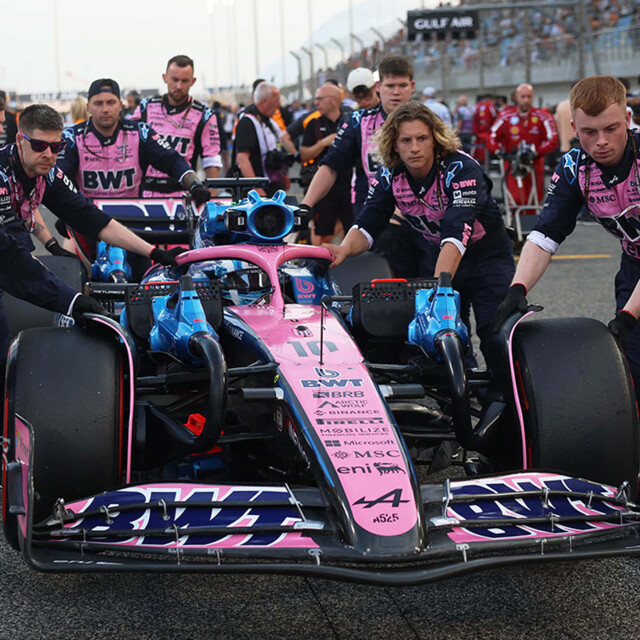 A focused Alpin/BWT racing team maneuvers their vibrant pink and blue Formula 1 car into position on the grid.