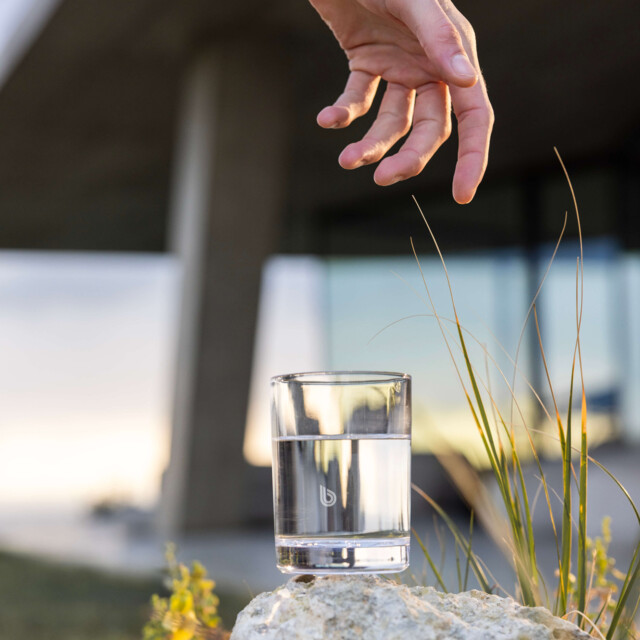 A clear glass of BWT pearlwater rests on a rocky surface surrounded by small plants, with a hand extending above as if to pick it up. 